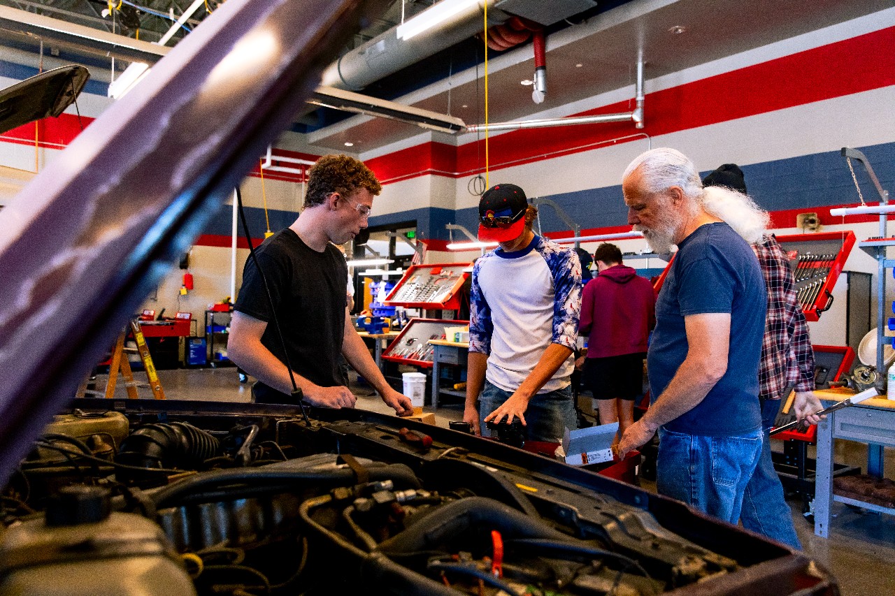 Two students inspect a car engine as their teacher observes.